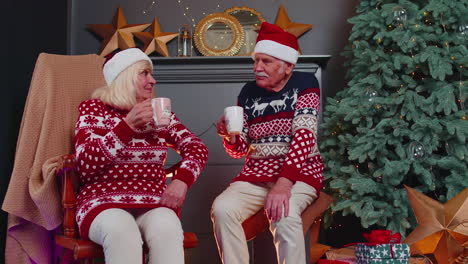 senior old wife and husband talking, drinking tea near glowing xmas tree on christmas in home room