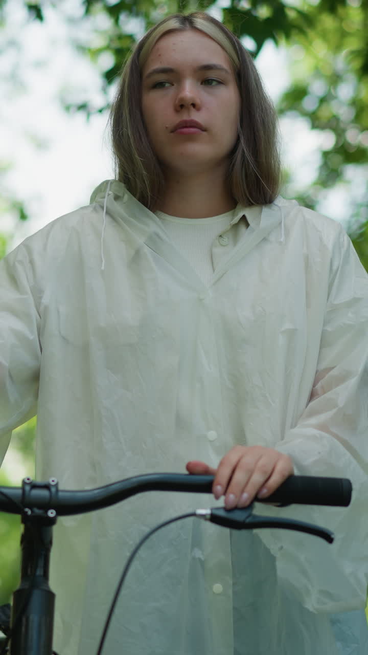 joven con impermeable translúcido se detiene mientras sostiene su bicicleta, quita la mano del manillar, y toma una respiración profunda, mirando perdido en el pensamiento, rodeado de vegetación exuberante y la suave luz del sol