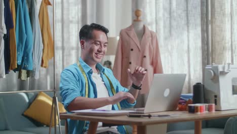 幸せなアジア人男性デザイナーがラップトップで作業しスタジオで服をデザインしながら祝っています