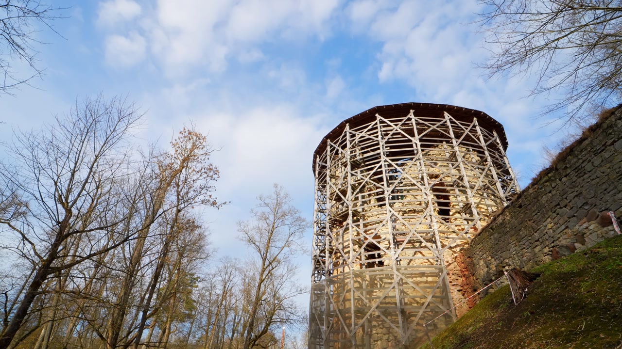Historic round tower wrapped in structural scaffolding on a sunlit hillside, with bare trees and early spring terrain showing restoration and heritage preservation work.