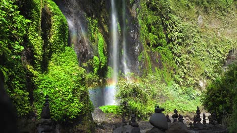 mojones de roca apilados en la base de una cascada tropical que crea un arco iris en el cañón de una selva tropical mística, java oriental, panorámica en cámara lenta hacia arriba