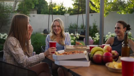 Three-girls-eat-pizza-communicate-laugh-during-lunch-at-a-table-in-the-courtyard-of-a-suburban-house