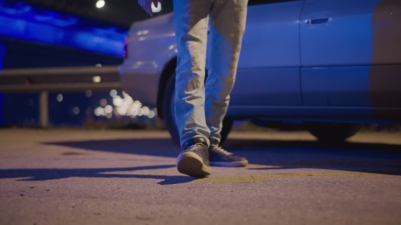 primer plano de una acera nocturna con los bajos y un pie cerca de una puerta abierta. Un conductor adulto de viajes compartidos en zapatillas y vaqueros se detiene junto a un sedán plateado bajo el resplandor azul neón y la luz de la calle. Disparo en ángulo bajo que revela un panel sombreado