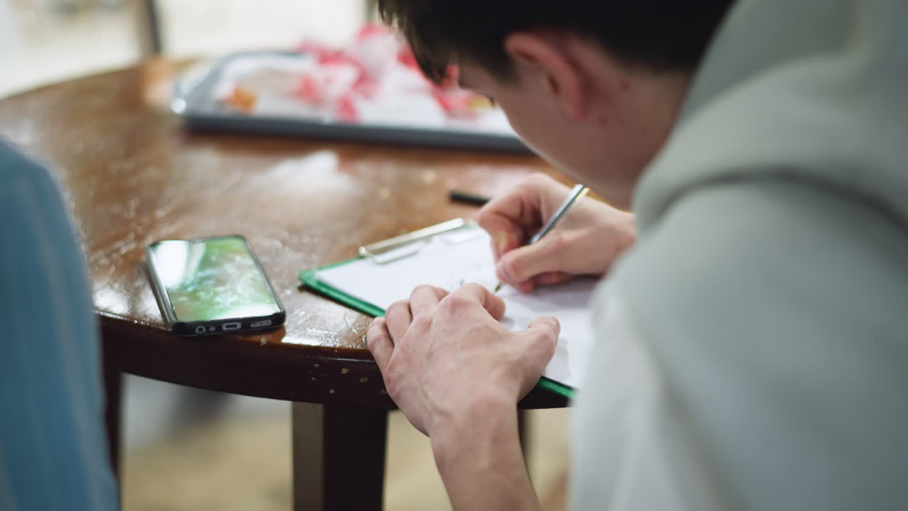 European looking boy guiding friend through sketching on printed image using pencil on paper while both lean over wooden desk bathed in soft natural light in study, facing bowl of snacks