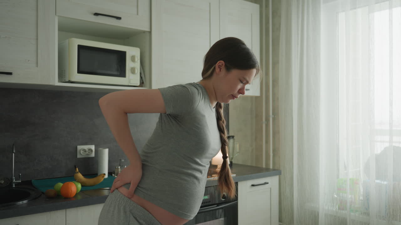 Pregnant caucasian woman holding lower back in kitchen with natural light, gray shirt and long braid, fruit on counter, tense expression, expectant mother coping with late pregnancy backache, gentle