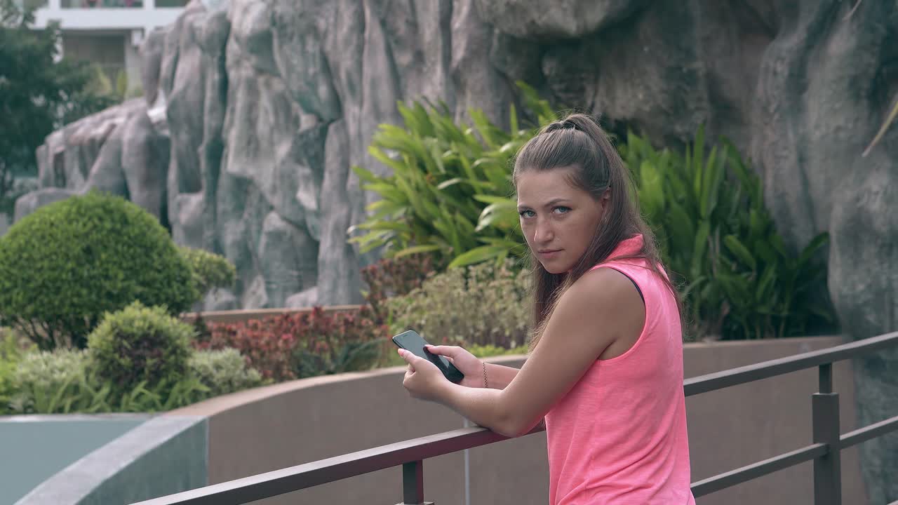 젊은 갈색 머리 소녀가 휴대 전화를 들고 밖에 서 있습니다.