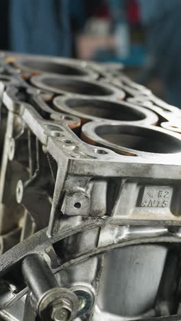 primer plano del motor del automóvil con detalles metálicos, pernos y cilindros, colocados en un soporte, personas con uniformes azules visibles en el fondo trabajando en un taller automotriz lleno de herramientas mecánicas
