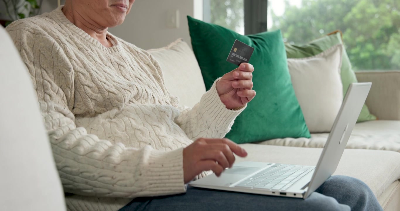 Anziano uomo asiatico con un maglione color crema a casa sul divano, digita sul laptop inserendo i dati della carta per pagare online.