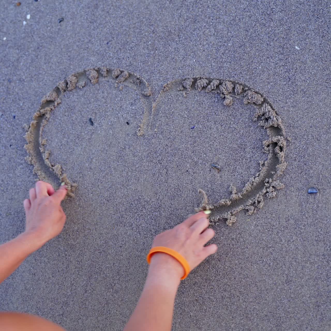 Girls hand draws a heart sign on a coastal sand, a holiday romance metaphor