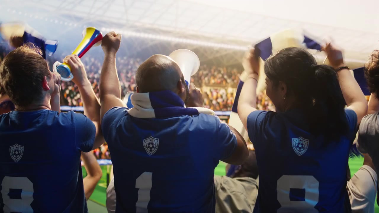 Aficionados al fútbol con camisetas azules haciendo sonar bocinas y megáfono en el estadio.