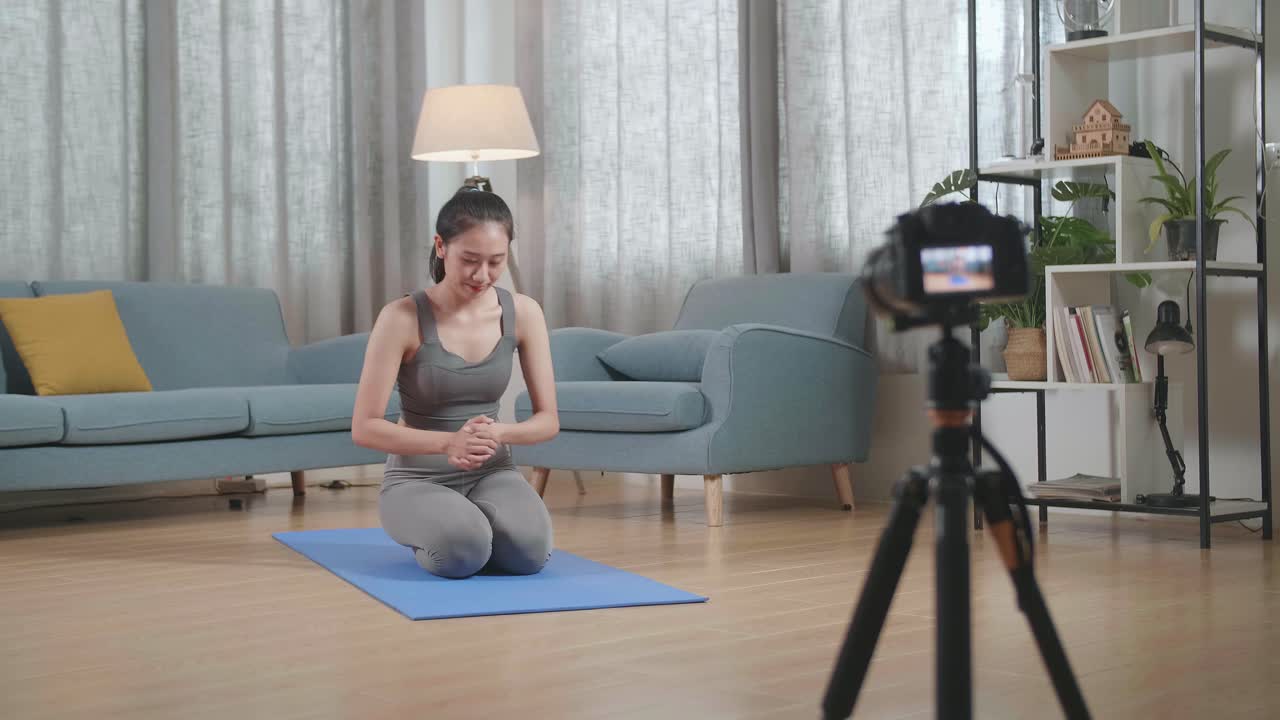 vista completa de joven entrenadora asiática mujer en ropa deportiva hablando a la cámara y haciendo yoga en la postura de tabla del antebrazo mientras graba el ejercicio de enseñanza en casa