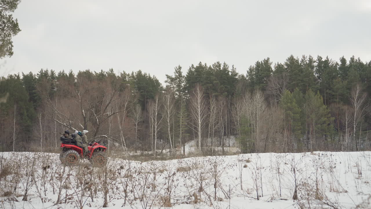 Un ATV in lontananza attraversa un campo innevato aperto, motociclisti solitari attraversano l'orizzonte delimitato da pini con spruzzi di neve e tracce fresche, esplorazione panoramica della natura selvaggia e serena avventura invernale in campagna remota.