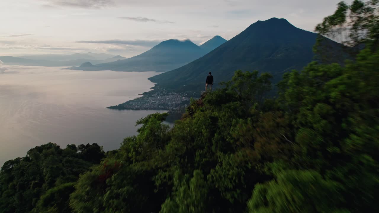 A spectacular drone shot captures a solitary figure standing on the iconic Indian's Nose viewpoint, overlooking the vast Lake Atitlán at sunrise