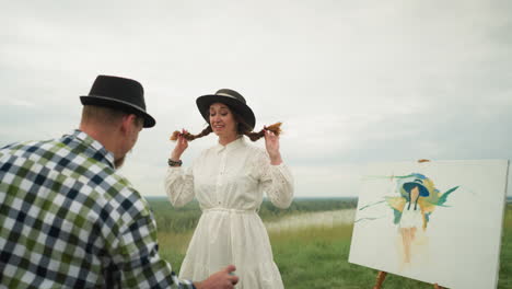 a man in a checkered shirt sprays mosquito repellent on a woman wearing a stylish hat and a white dress. they are standing in a lush grass field with a partially painted canvas in the background