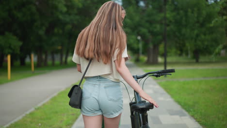 back view of young woman in jean shorts and crop top, with black handbag, walking alongside her bicycle, hand on seat, enjoying a stroll through green park path lined with trees and nature