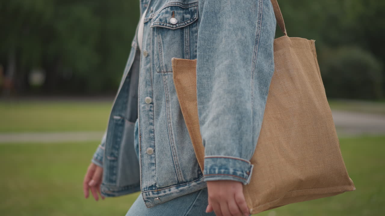 Borsa di paglia vista laterale, passeggiando nel parco, giacca di jeans e borsa intrecciata che ondeggiano, sfondo verde e passo disinvolto, atmosfera da mercato sostenibile, commissioni estive con contrasto di texture tra paglia e denim.