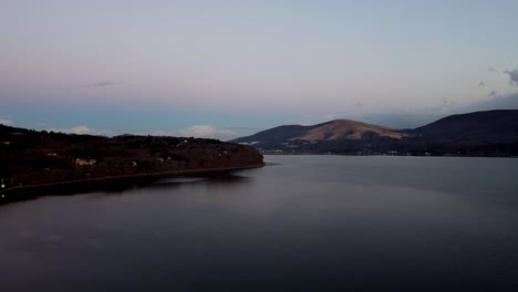 Tonos-Crepusculares-Sobre-Un-Lago-Tranquilo-Con-Colinas-Distantes-Y-Luces-Dispersas,-Ambiente-Nocturno-Sereno,-Vista-Aérea