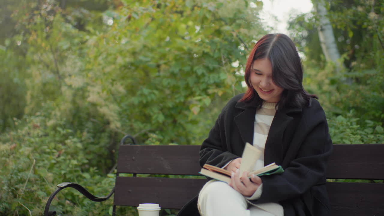 Female student seated on park bench outdoors, legs crossed, smiling while flipping book pages, paper cup by side, peaceful greenery around, study break mood, quiet reading moment with gentle laughter