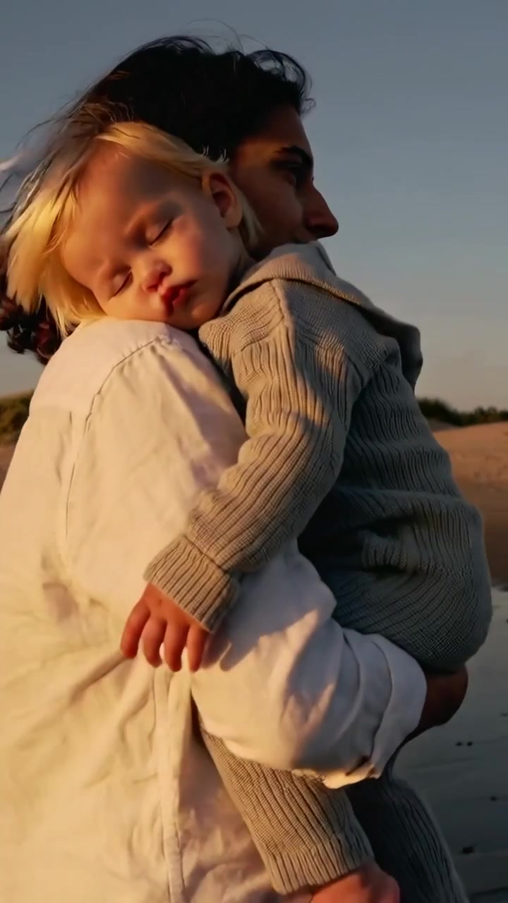 Video verticale: Papà che tiene in braccio un bambino per calmarlo, si sistema sulla riva al tramonto con un completo in maglia e una camicia chiara