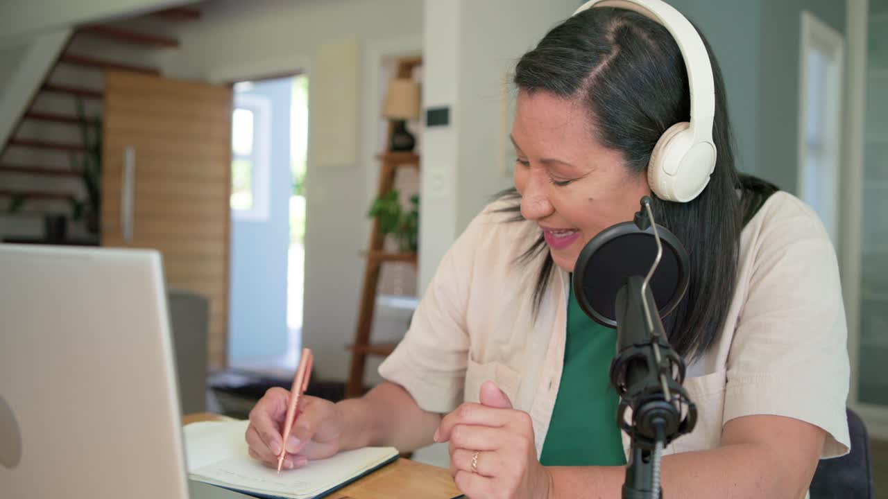 Mujer madura iniciando un podcast, hablando por el micrófono, gesticulando y tomando notas en el portátil en casa.