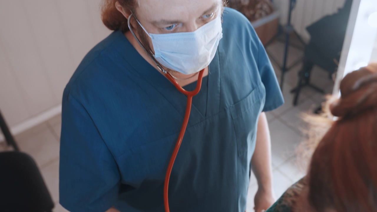 Visita domiciliare di un paramedico (vista dall'alto) con stetoscopio, un medico bianco controlla la spalla del paziente con un maglione verde, l'interazione mostra intesa e valutazione professionale in un interno domestico, atmosfera di cura d'urgenza.