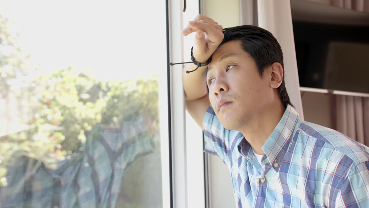 Pensive asian man in plaid shirt looking out window, reflecting on thoughts, copy space
