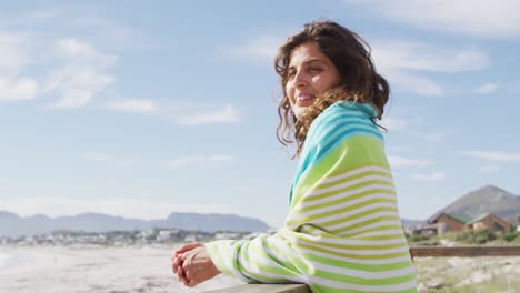 Happy-mixed-race-woman-standing-by-the-sea-with-blanket-over-shoulders-enjoying-the-view-and-smiling