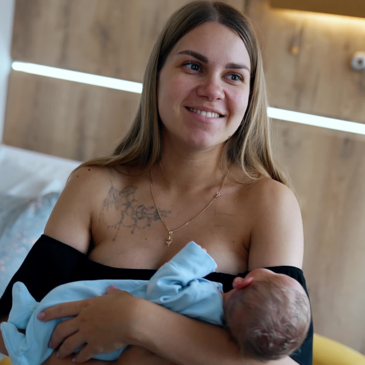Happy smiling Caucasian woman in black dress with bare shoulders holding a baby. Smiling mother waving her tiny newborn son