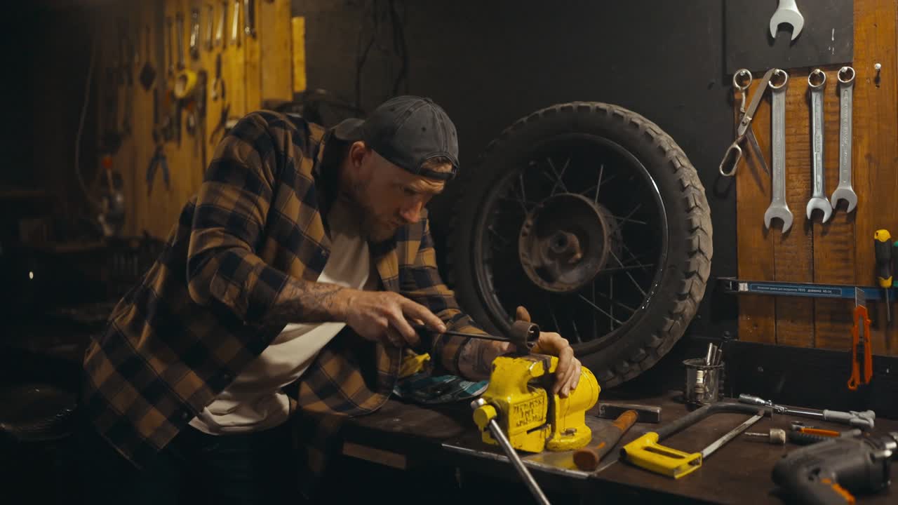 Focused mechanic with a beard in a plaid shirt works with a file on a workbench in his repair shop studio