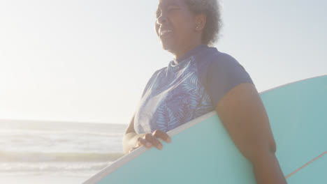 una mujer afroamericana mayor feliz caminando y sosteniendo una tabla de surf en la playa, en cámara lenta