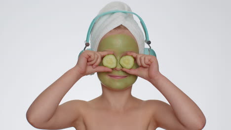 smiling child girl moisturizing green cucumber mask on face listening to music on headphones dancing