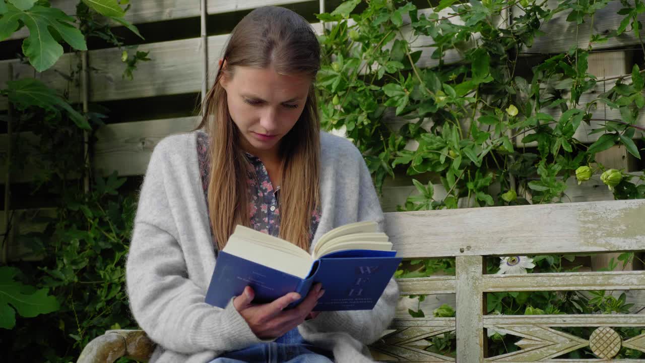 chica con cabello largo se sienta leyendo un libro en un banco de jardín al aire libre