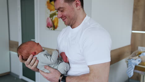 handsome caucasian man smiles to a tiny baby he's holding in hands. father dandles his newborn in hospital ward.