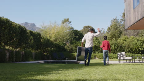 indian father and son walking together in backyard near swimming pool and chairs, copy space