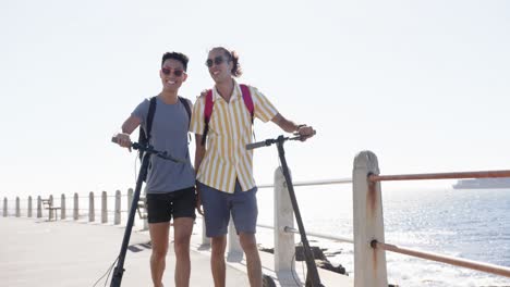 happy diverse gay male couple walking with scooters at promenade by the sea, slow motion