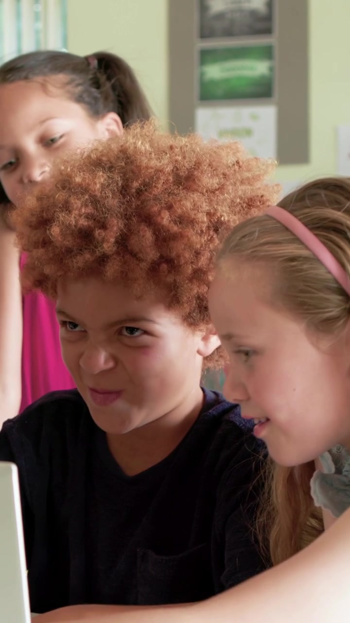 Vertical video: Reaching curly-haired boy operating laptop in classroom, classmates watching screen