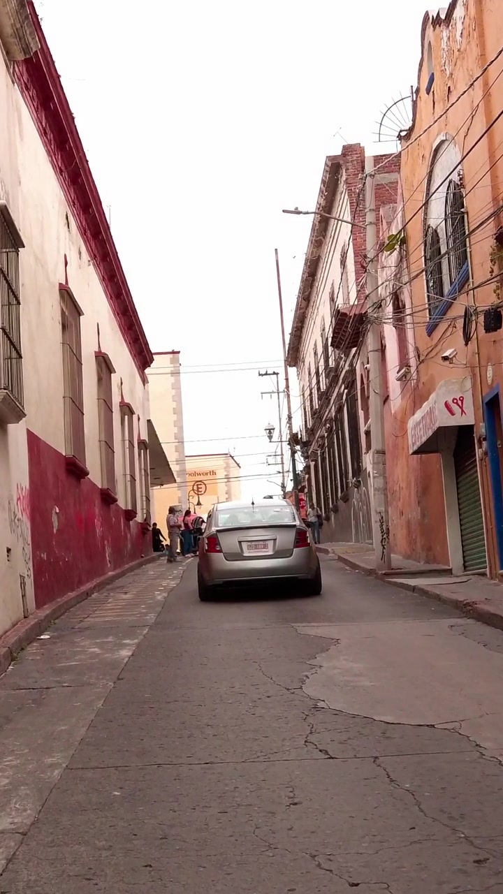 Stretta via urbana fiancheggiata da edifici storici, balconi e cavi di servizio aerei, con un'auto parcheggiata, pedoni e pavimentazione fessurata che creano un'autentica scena di quartiere cittadino.