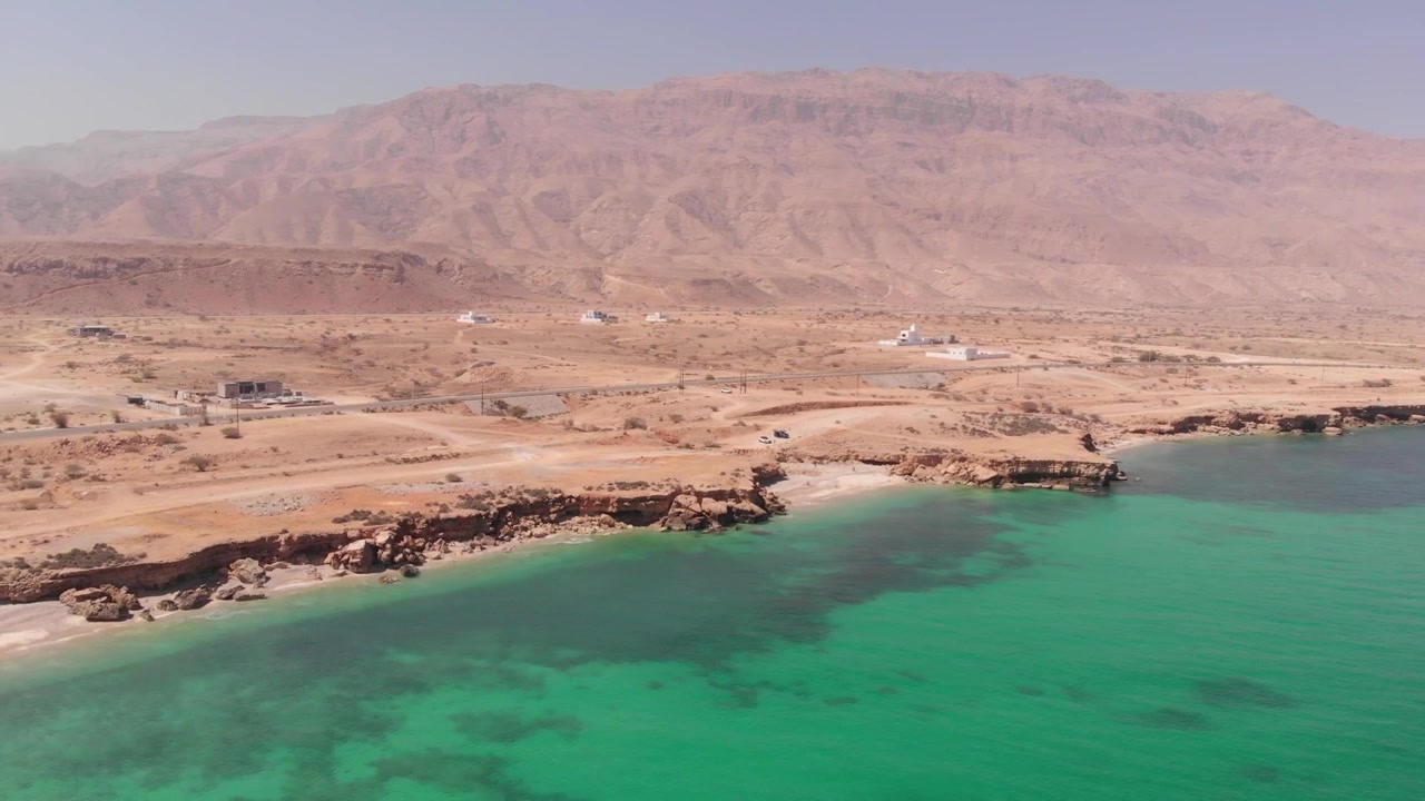 Ripresa aerea con drone dall'alto di una strada costiera tortuosa lungo le onde turchesi dell'oceano e le aspre scogliere desertiche dell'Oman. Mette in mostra il drammatico contrasto tra paesaggio arido e mare.