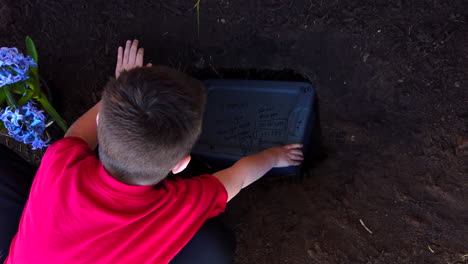 Un-Niño-Colocando-La-Caja-De-Plástico-Del-Cuerpo-De-Una-Mascota-Familiar-Muerta-En-Un-Hoyo-Recientemente-Excavado-En-Un-Jardín-Familiar-Para-Enterrarlos-Allí-Y-Tener-Su-Tumba-En-El-Jardín