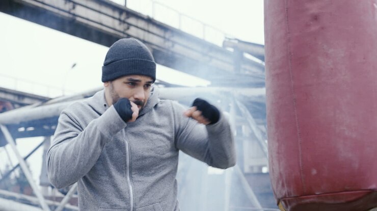Vista laterale di un uomo caucasico in abbigliamento sportivo che colpisce un sacco da boxe all'aperto, in una fabbrica abbandonata, in una mattinata nuvolosa.