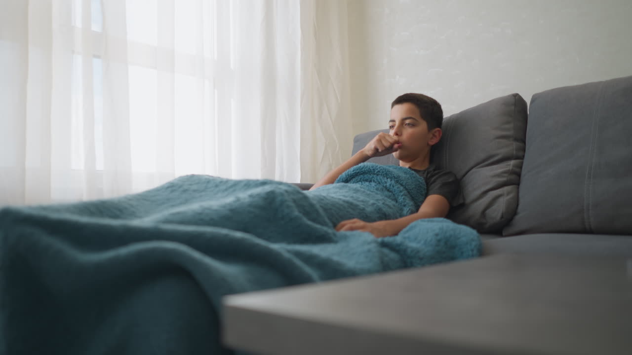 Little kid feeling cold lies on sofa near window, wrapped in thick blanket as he coughs and covers mouth with hand, showing signs of illness and fatigue in calm softly lit modern living space