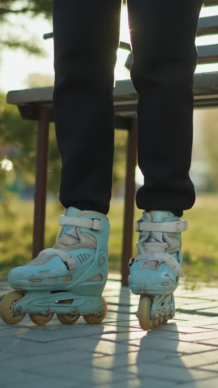A person dressed in black trousers is standing near a park bench, ready to skate with rollerblades on their feet. A pair of black boots is placed on the ground beside them