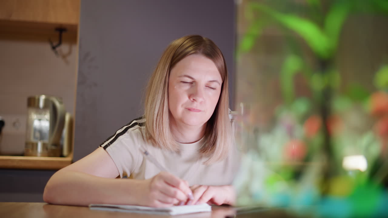 Woman sitting at desk writing in notebook with pen, focused expression showing concentration and mindfulness, in calm home environment with fish tank adding relaxing atmosphere