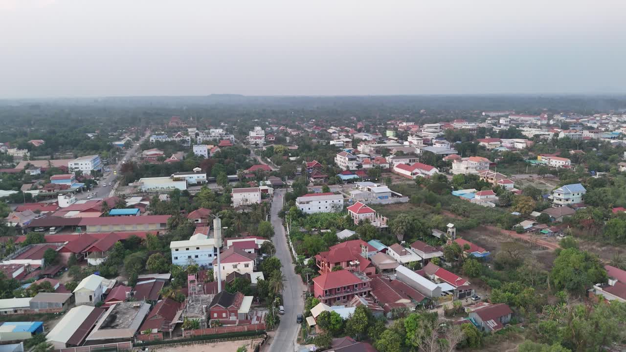 This aerial shot captures a peaceful Southeast Asian town, possibly Siem Reap, under soft, dusky light. Low to medium-rise buildings with varied roofs are interspersed with lush greenery