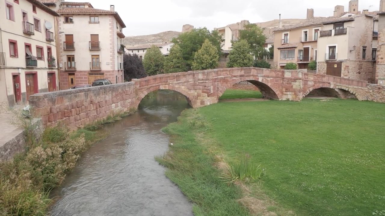 Puente de piedra histórico sobre un pequeño río junto a casas tradicionales en un pueblo español.