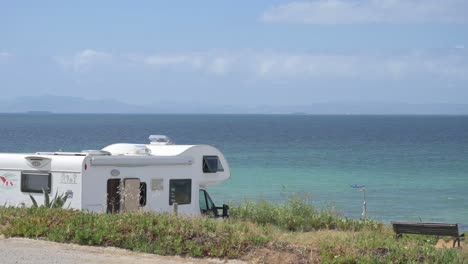 a white caravan is parked by the calm sea with mountains in the distance. blue sky and clear water