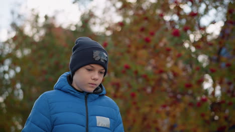 kid wearing black beanie with patch and blue jacket strolling with head bowed thoughtfully, surrounded by blurred autumn foliage and vibrant colors