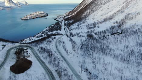 Vista-De-Drones-Sobre-Las-Montañas-De-Tromso-En-Invierno-Llenas-De-Nieve-Mostrando-Un-Camino-Serpenteante-Con-Una-Pequeña-Isla-Con-Un-Pueblo-En-Husoy-Junto-Al-Mar