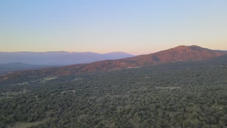 Vuelo-De-Drones-Al-Atardecer-En-La-Hora-Dorada-Sobre-Un-Bosque-Con-Vistas-A-Una-Colina-Con-Un-Fondo-De-Montañas