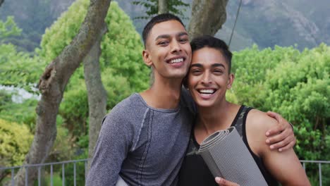 Portrait-of-happy-mixed-race-gay-male-couple-standing-in-garden-embracing-and-laughing